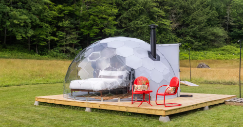 A glamping dome with a bed inside, surrounded in a field by red lawn chairs and flanked by the forest.