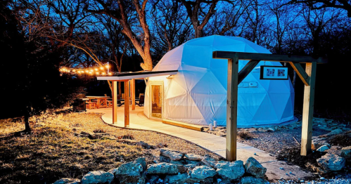 A glamping dome glows softly at twilight, with string lights illuminating a nearby outdoor dining area.