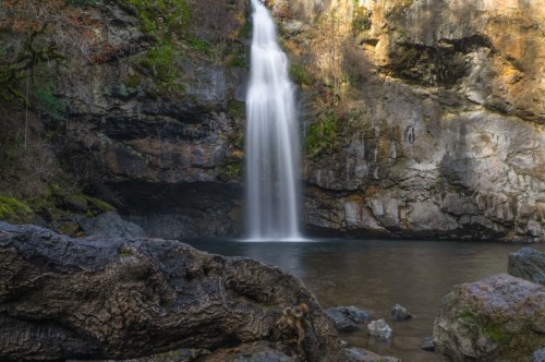 Rocks border a thin waterfall with a swimming hole below.