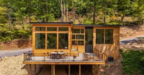 Modern wood cabin with large windows and a balcony overlooking trees. Features a flat roof and outdoor dining set.