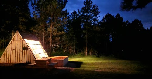 Warmly lit A-frame SD cabin glows at night. A picnic table sits on the grass, surrounded by silhouetted trees.