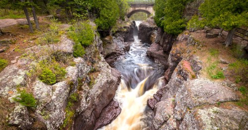 A narrow river rushes under an arched bridge into a narrow, rock-walled gorge topped with green trees and bushes.