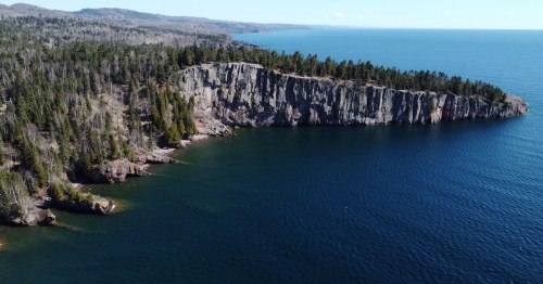 Evergreens line the gray rocky shore of Lake Superior, which reflects the clear blue sky.