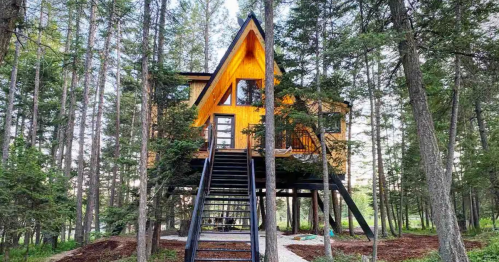 A-frame treehouse cabin on stilts with a metal staircase in a dense Montana forest, close to Glacier NP.