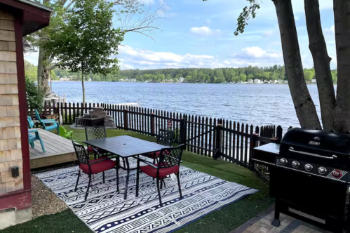 The fenced-in backyard at the Little Lake House Airbnb property in Derry, New Hampshire. The backyard overlooks a lake. A table with four chairs and a grill is pictured.