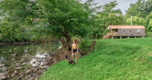 a woman and her dog standing on grass near a waterfront cabin in west virginia