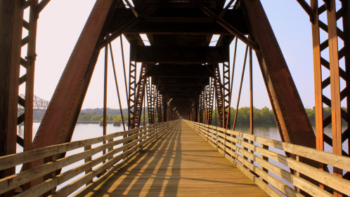 A wooden pedestrian walkway extends through the center of a steel railroad bridge spanning the Tennessee River on a summer day.