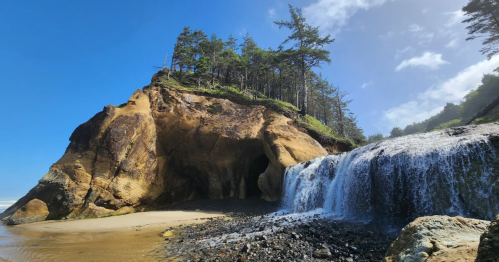 An oceanfront waterfall pours from a local stream onto the beach beside a rugged outcrop of rock and trees.