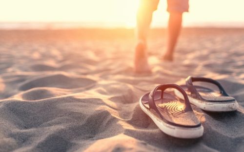 A person walks barefoot on the beach, leaving flip-flops in the sand as the sun sets in the background.