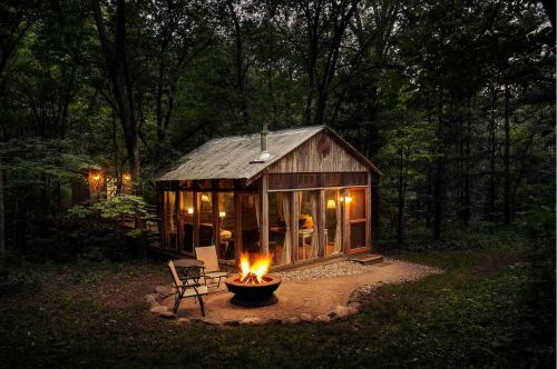 A cabin in the woods with a fire lit in the pit.