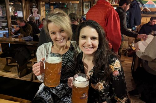 Two women holding large beers while sitting on a bench.