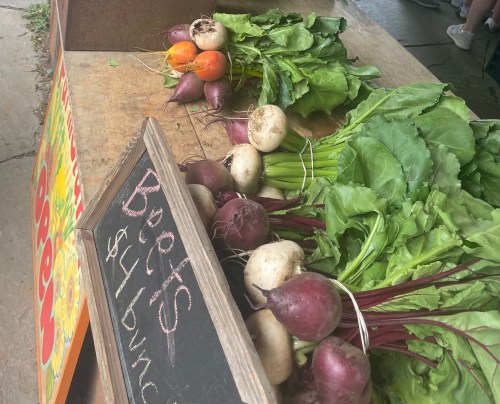 A table at a farmstand full of fresh red beets, with lush green leaves. There is also a sign displaying their price.