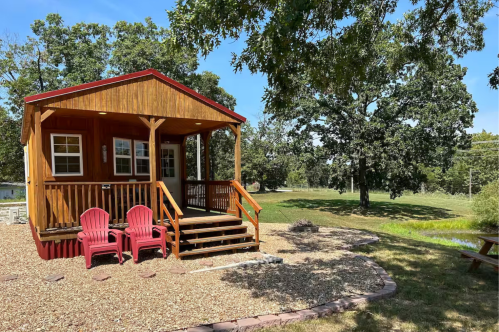 Two red Adirondack chairs sit in front of a rustic wood cabin by a pond.