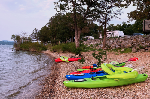 A row of colorful kayaks line a lake shore with RV campers seen in the distance.