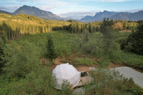 A geodesic dome is set up in the middle of the forest, surrounded by stunning mountain scenery.