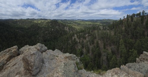 Rocky overlook above a forested canyon with rolling Black Hills and a partly cloudy sky in the distance.