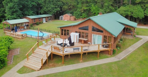 a large cabin with a deck and a pool sitting on a large grassy yard with forest trees behind