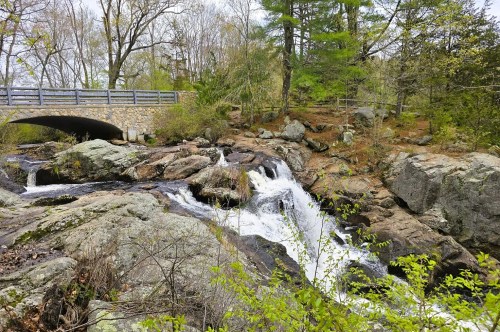 A flowing waterfall cascading over tiered rocks in a wooded area with a stone bridge above it