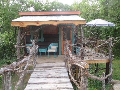 A wooden bridge leads to a small treehouse, with a wraparound porch and quirky teal details. The house is surrounded by trees.
