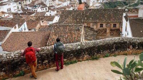 Two hikers stand on a stone wall overlooking a picturesque village nestled in a valley surrounded by mountains.