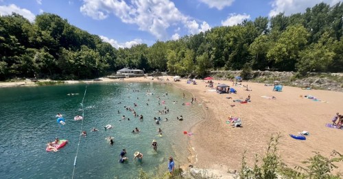 A quarry swimming hole in Indiana featuring a crowded sandy beach, surrounded by heavy forestry.