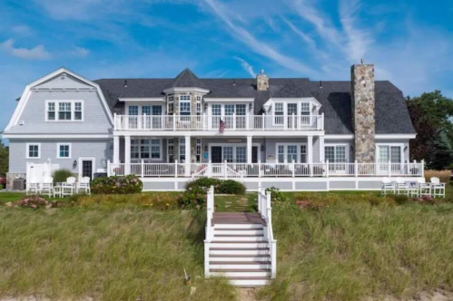 An exterior shot of The back of the Beach Estate VRBO property in Mattapoisett, Massachusetts. The large beach home has a double porch and a set off stairs leading out to the beach.