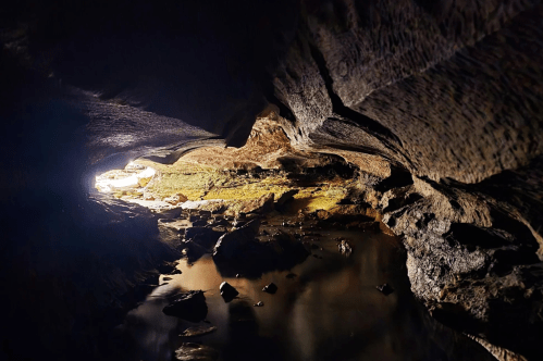 An image taken in a cave/sinkhole shows dark rocks framing the view of a waterway and an opening to the sunlight outside.