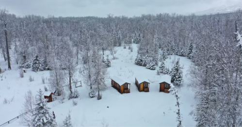 Three small cabins are seen from above on a snowy day, their roofs covered in fresh snow.