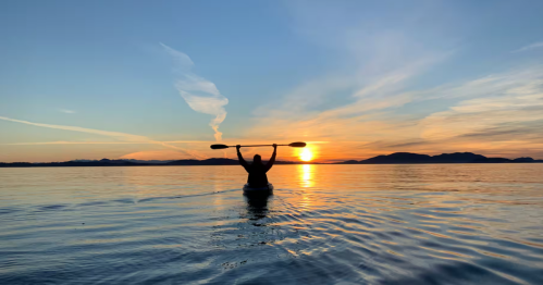 At a stunning golden hour sunset over the water, a kayaker lifts his paddle above his head.