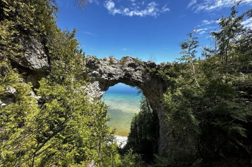 An arched rock formation in a forest with a lake in the background.