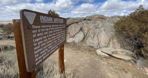 A sign explaining the history of the Indian Bathtubs in rural Wyoming stands next to a dirt trail and large rocks.