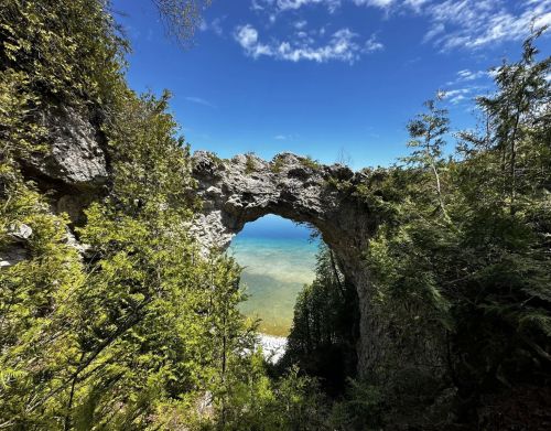 An arched rock formation in a forest with a lake in the background.
