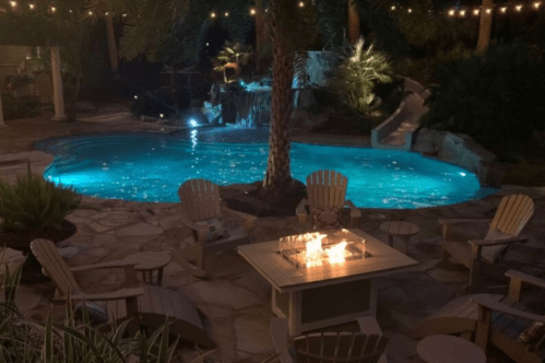 Pool with a slide and waterfall at night with a neighboring firepit and chairs