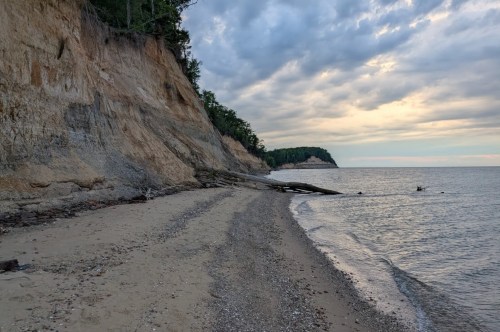 The sun is setting on a beach with tall cliffs and a few pieces of timber washed up on shore.