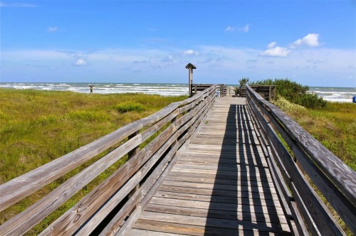 Views of a wooden boardwalk leading to a beach, surrounded by grassland along a coastal hiking trail in Texas.
