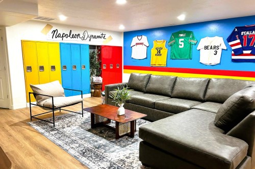 A Napoleon Dynamite-themed Airbnb in Utah with yellow, blue, and red lockers, a blue wall with various sports jerseys, and a sitting area with sofas and a chair.