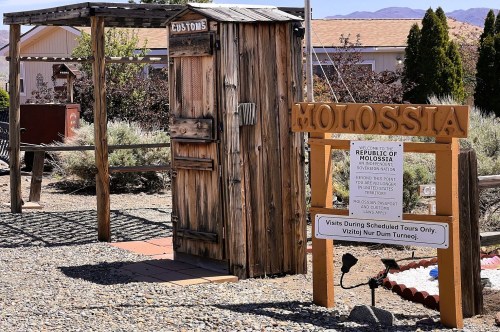 A wooden sign for Molossia next to a small wooden customs building with trees and houses in the background, inside a quirky micronation in Nevada.