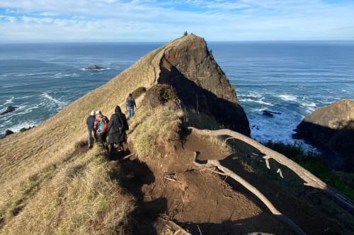 A sloping rock juts upward with a trail and hikers leading to it, the ocean is in the back with a cloudy blue sky overhead.