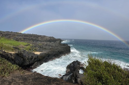 A dark rocky shoreline with a churning ocean has a low-hanging rainbow curving over it.