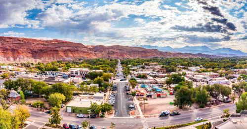 A panoramic view of Moab, Utah, along Main Street, surrounded by red rock mountains and greenery, various buildings, and parked cars.