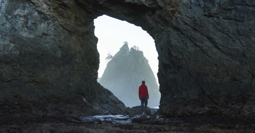 A man in a red jacket stands beneath an arch at Rialto Beach in Forks, Washington.