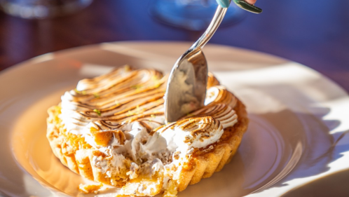 Hand cuts into a coconut tart with toasted meringue. Tart on white plate on a wooden table.
