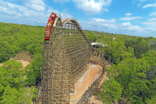 A giant wooden roller coaster rises above a green forest that extends to the horizon.