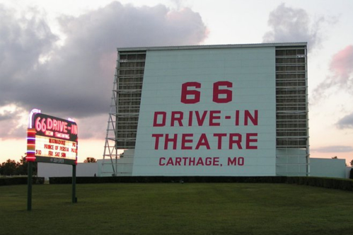 A large billboard has red text with the name of the 66 Drive-In Theatre behind a neon marquee advertising film titles.