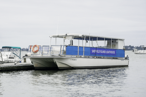 White catamaran ferry, "Sheffield Island Lighthouse," docked in calm water on an overcast day.