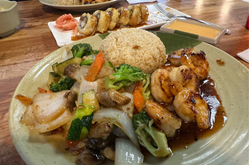 A hibachi plate shows grilled vegetables and shrimp beside a pile of rice with a sushi roll in the distance.