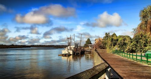 View of a coastal town in Georgia with a boardwalk to the right and a shrimp boat to the left, featuring blue skies and clouds.