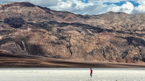 A lone man in red walks along the white sands of Death Valley National Park, the mountains looming in the distance.