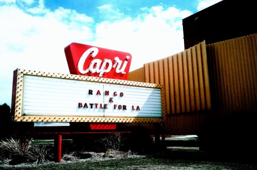 A large red and white marquee sign for Capri Drive-In movie theater in Michigan, next to a dark-colored wall, surrounded by shrubs and a grassy surface.