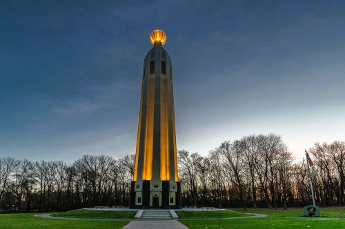 A large glowing tower with an illuminated light bulb on top, set inside a grassy area with paved pathways and surrounded by a wooded area in the background, at an iconic roadside attraction in New Jersey.
